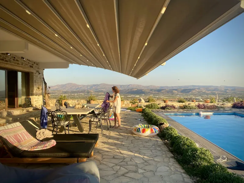 Store banne à toile beige déployé au-dessus d’une terrasse en pierre, avec piscine et collines à l’horizon