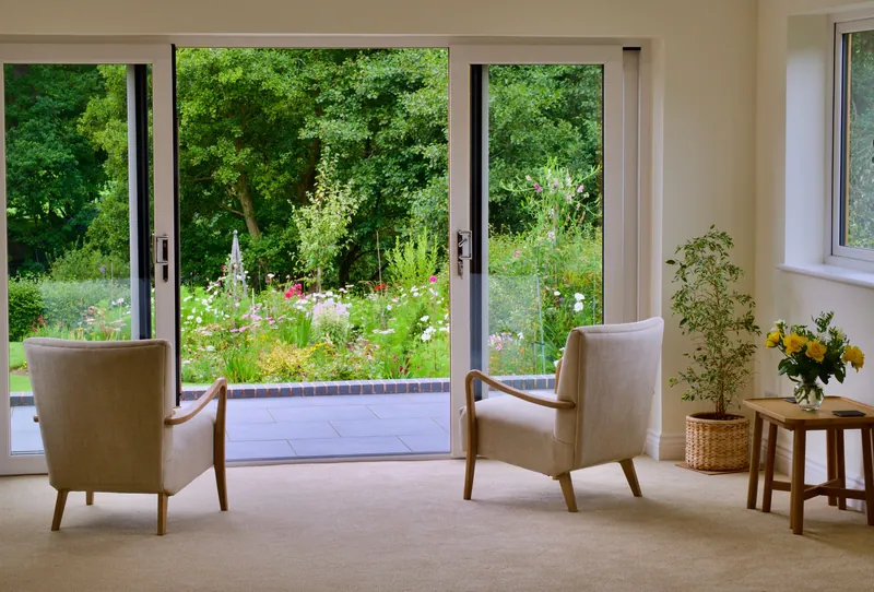 Baie vitrée coulissante blanche ouverte sur un jardin fleuri, deux fauteuils dans le salon