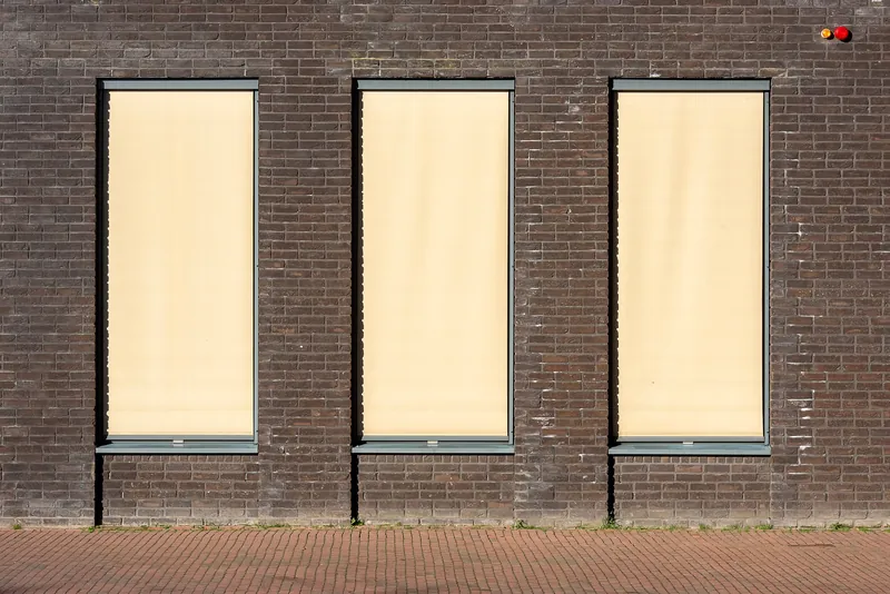 Trois fenêtres d’une façade en briques couvertes par des stores extérieurs verticaux à toile beige