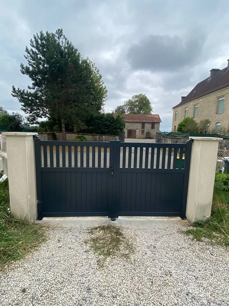 Portail battant anthracite à 2 vantaux, ajouré en partie haute, entre deux piliers beiges devant une cour