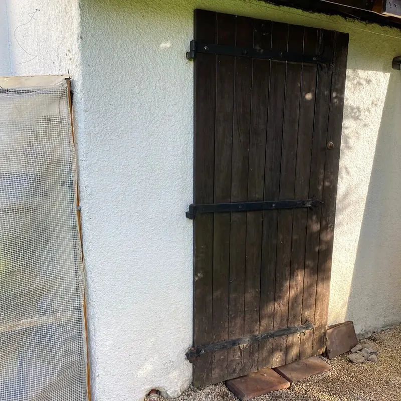 Avant : Ancienne porte en planches de bois sombre sur le mur blanc de l’abri, avant son remplacement