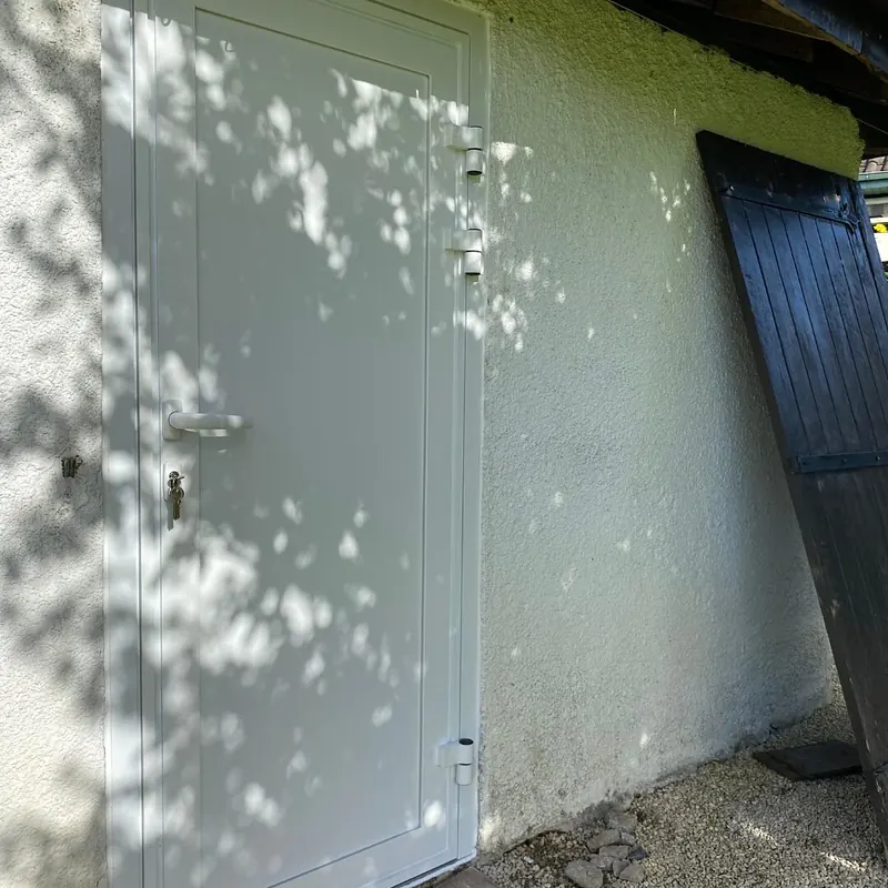 Après : Porte pleine claire posée sur le mur blanc d’un abri, l’ancienne porte en bois appuyée à côté