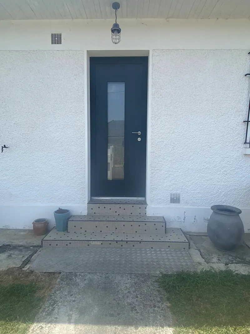 Porte d’entrée anthracite à vitrage vertical étroit et poignée inox, en haut d’un perron en mosaïque