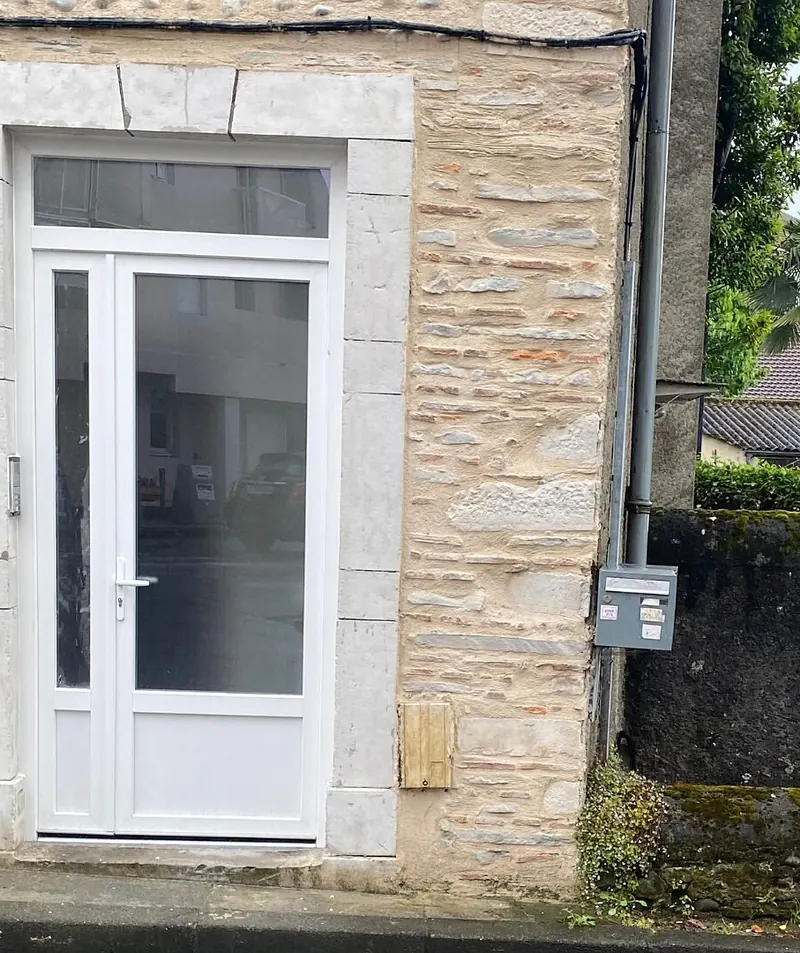 Après : Porte d’entrée PVC blanche vitrée, avec partie latérale étroite et imposte vitrée, posée dans un encadrement en pierre de taille sur une façade en galets et moellons